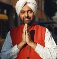Smiling man in traditional attire and turban with a greeting pose outdoors.